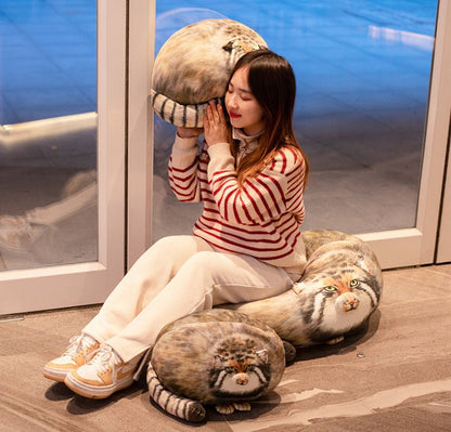 lady sitting on a big size fat cat plushie and holding another one and these plushies are inspired by pallas' cat