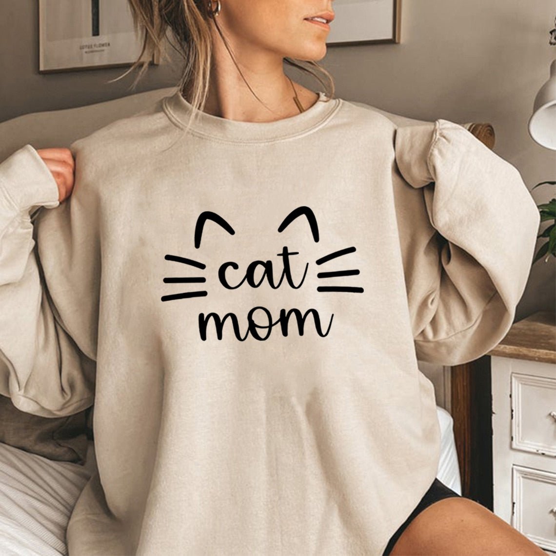 a woman wearing a beige color womens cat sweatshirts with the word cat mom on it
